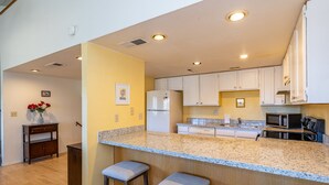 A kitchen with a granite countertop, white cabinets, a refrigerator, a microwave, and two bar stools.