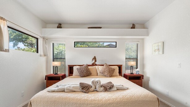 A neatly made bed with a quilted cover, two bedside tables with lamps, a window with blinds, and a small shelf with decorative items. - Waterfall, Grapevines and Hot Tub with Views - Nice House Too! (Sedona)