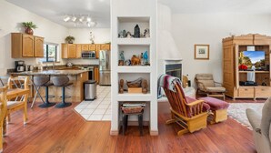 A modern kitchen with wooden cabinets, a central island, and bar stools. There is a white shelving unit with decorative items and a seating area with a cushioned chair and ottoman. - Waterfall, Grapevines and Hot Tub with Views - Nice House Too! (Sedona)