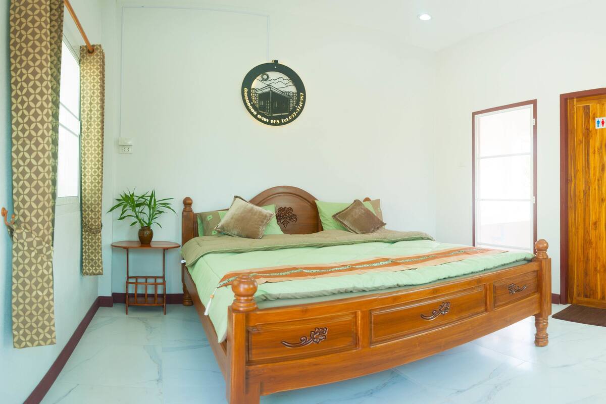 Un dormitorio con una cama de madera, una mesita con una planta, una puerta con un cartel y un refrigerador.