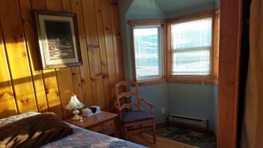 Un dormitorio con cama, una silla, mesita de noche, lámpara y ventana con persianas.