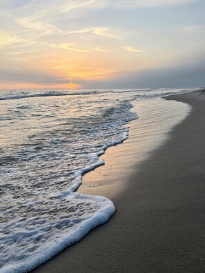 Una spiaggia con onde che lambiscono dolcemente la riva, cielo sereno e tramonto in lontananza.