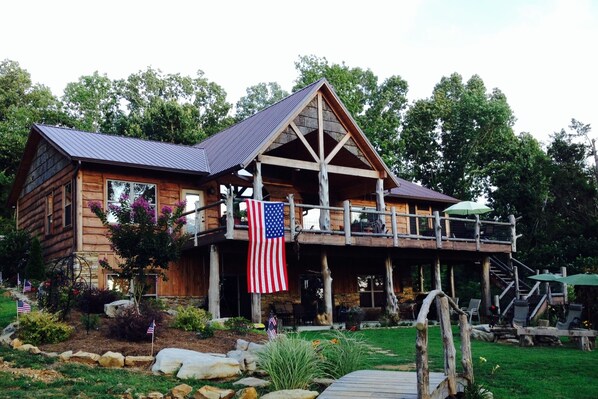 A wooden cabin with a balcony, an American flag, and a stone foundation.