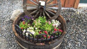 A rustic wooden barrel planter filled with colorful flowers, placed on a gravel surface. - Your "Home Away From Home"-Rustic Alaskan Home (Soldotna)