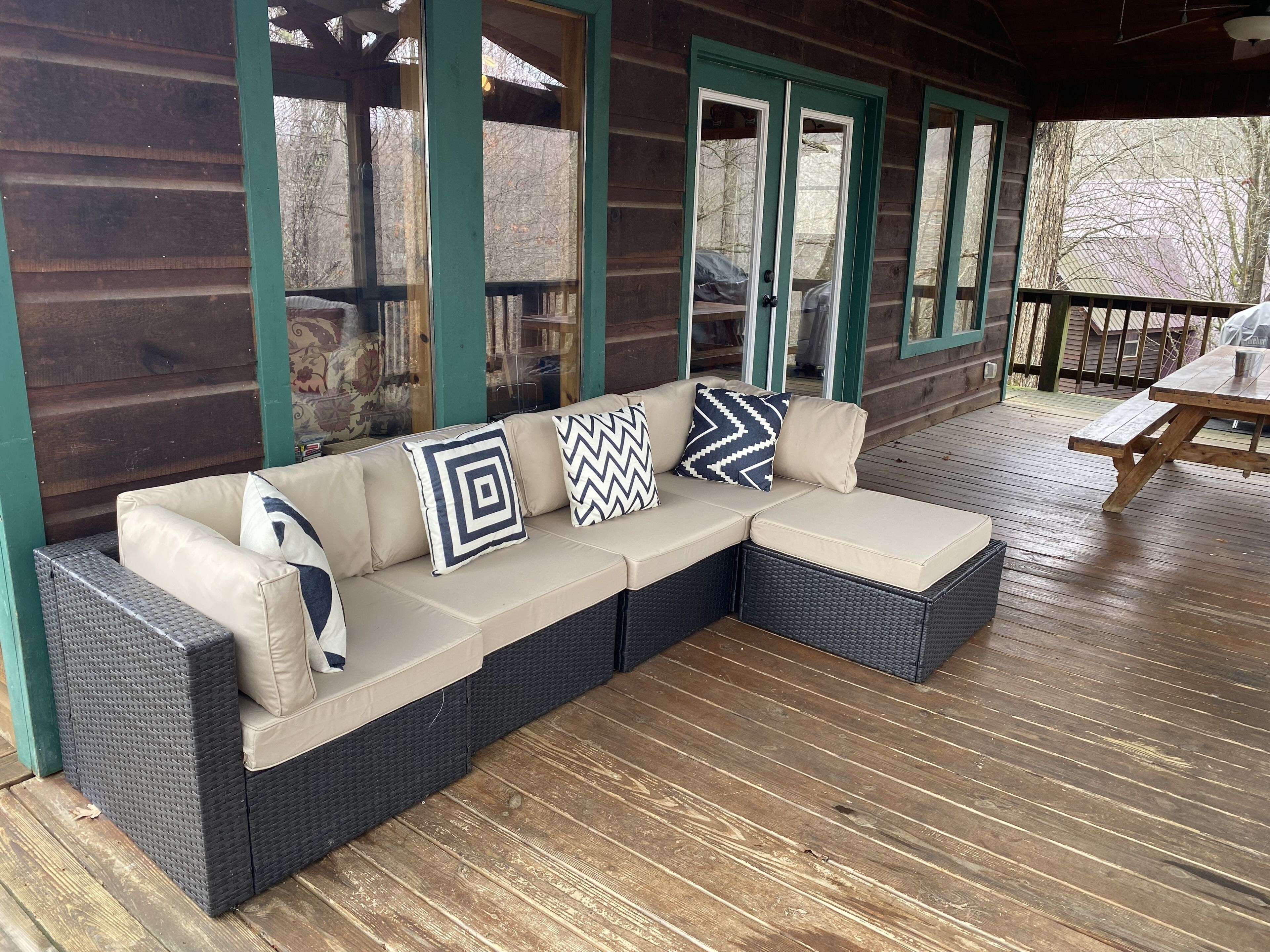Un espace extérieur aménagé, abrité et doté d’un canapé en osier et de coussins, d’une terrasse en bois et d’une vue sur les arbres à travers les fenêtres.