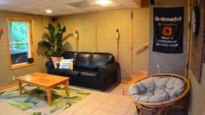 Een kamer met een zwarte leren bank, een houten bijzettafeltje, een rieten stoel, een grote potplant en een spandoek aan de muur.