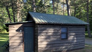 A small wooden cabin with a dark door and a window, surrounded by trees.