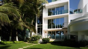 Un bâtiment moderne à plusieurs étages, doté de grandes fenêtres en verre, d’une façade blanche et d’un jardin bien entretenu avec des palmiers et un chemin menant à l’entrée.