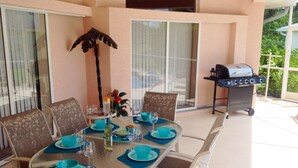 An outdoor dining area with a round table set for four, surrounded by white chairs. A grill is visible in the background.