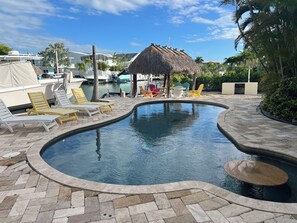 Uma piscina com espreguiçadeiras e uma estrutura com telhado de palha, cercada por palmeiras e barcos.