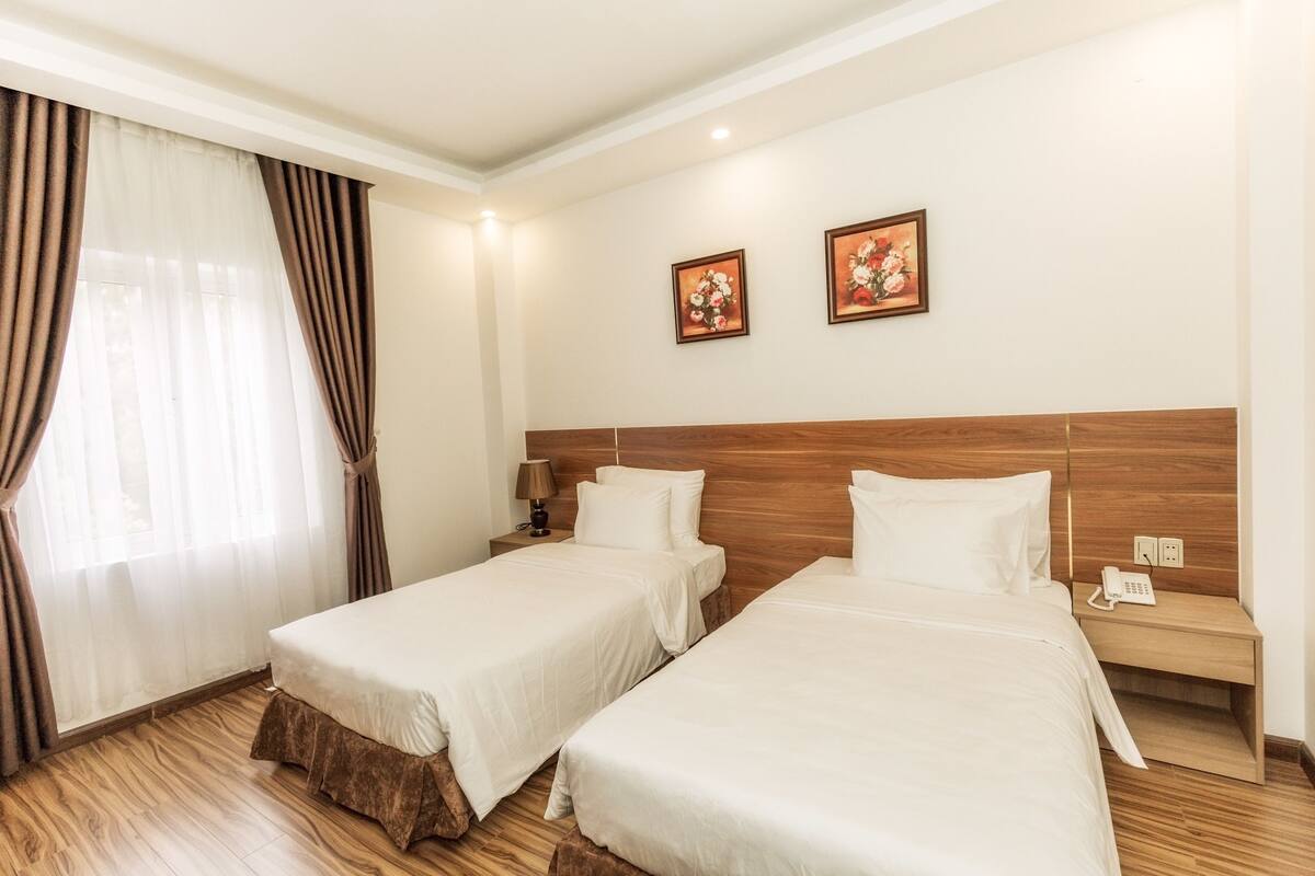 A hotel room with two single beds, a wooden headboard, a nightstand with a lamp, and two framed floral pictures on the wall.