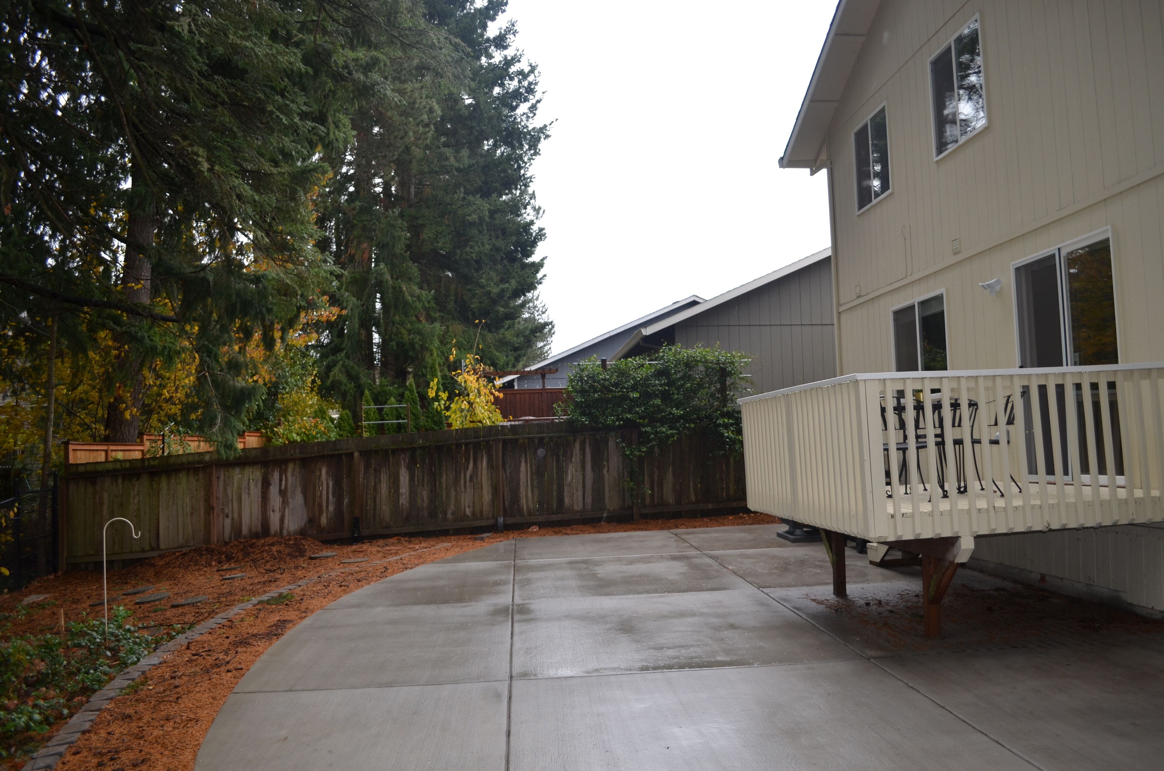 A residential backyard with a concrete patio, a wooden fence, and a white deck.