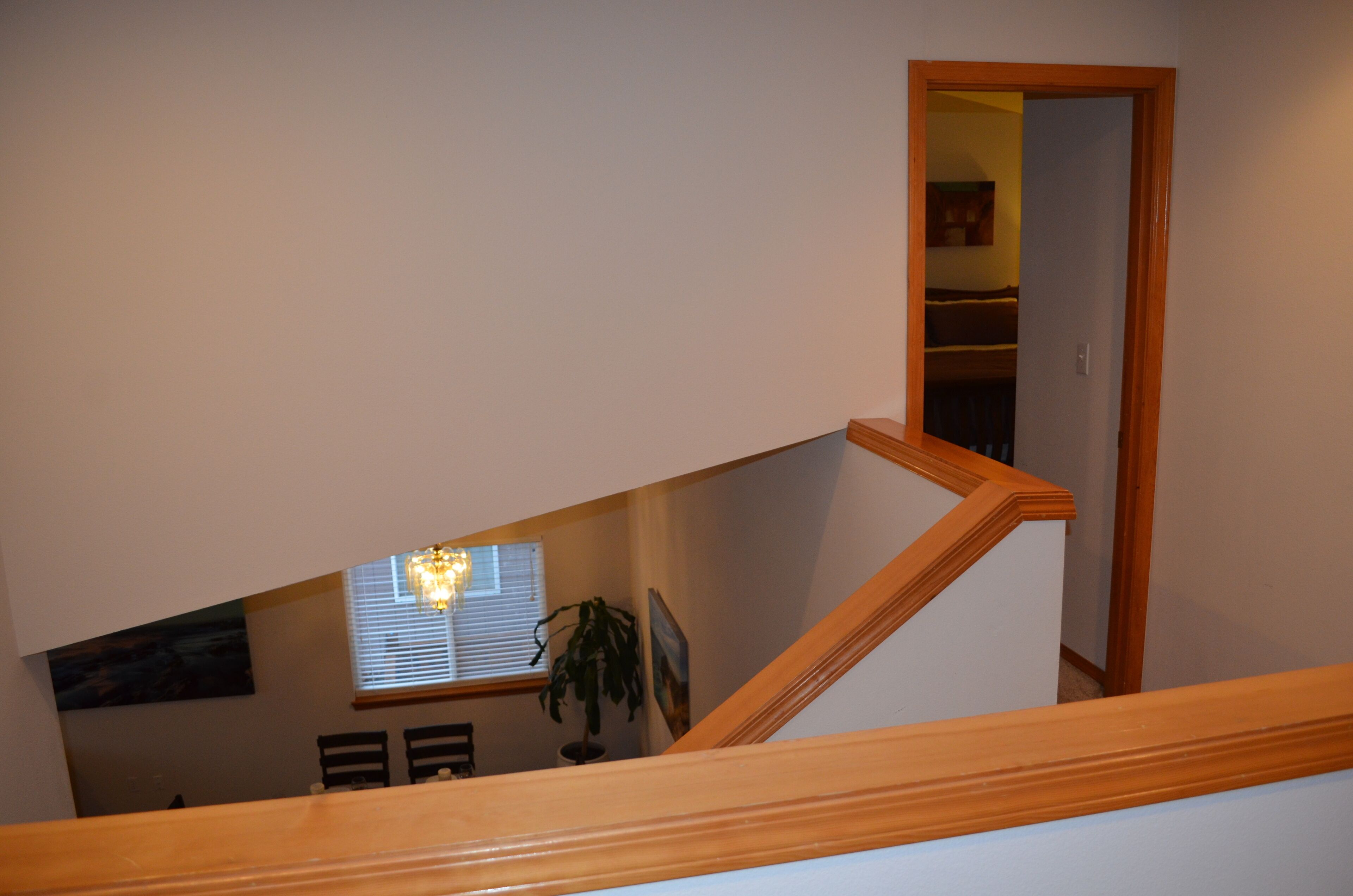 A staircase with wooden railings leading to a second-floor area with a dining table, chairs, a potted plant, and a window with blinds.