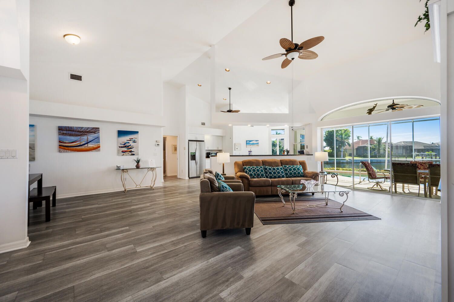 Ein geräumiges Wohnzimmer mit einem braunen Sofa, einem Glastisch und einem Essbereich mit Tisch und Stühlen. Im Hintergrund ist eine Küche mit weißen Schränken und einem Kühlschrank zu sehen. Große Fenster bieten einen Blick ins Freie.