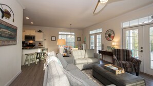 A spacious living area with a large sectional sofa, a dark armchair, and a dining table with chairs. A kitchen is visible in the background, and there is a ceiling fan above. - Immaculate, 4 Bedrooms/4 Bath, w/ Pool, Exterior Elevator and Golf Cart (Emerald Isle)