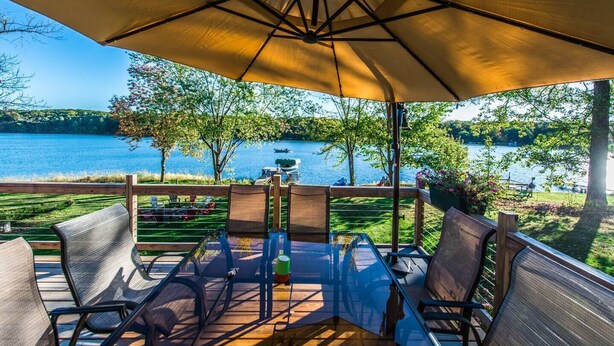 Eine Terrasse mit Sitzgelegenheit und einem großen Sonnenschirm mit Blick auf einen See, auf dem Boote und Bäume zu sehen sind.