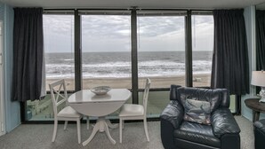 A room with a large window overlooking the beach, a white table with chairs, and a black leather armchair. - Oceanfront & Pool, on the Boardwalk at 37th Street (Virginia Beach)