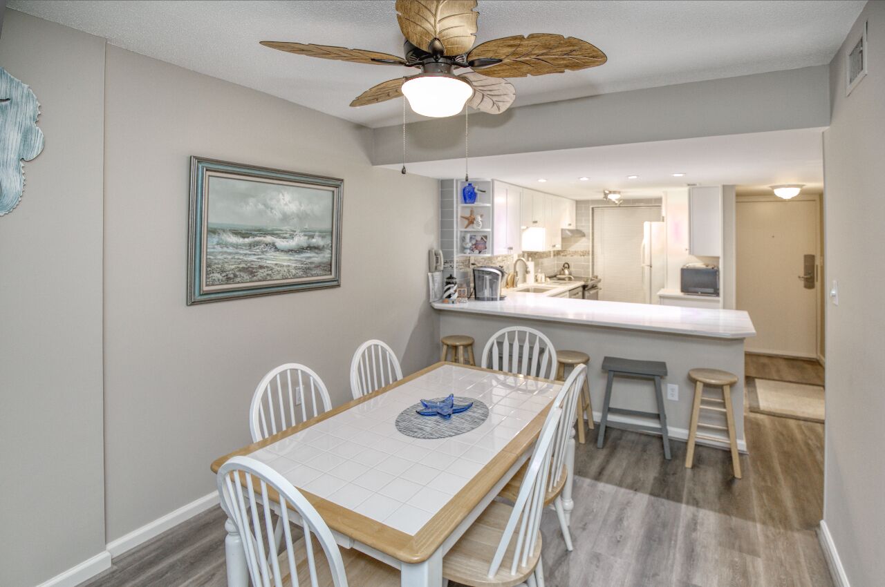 A dining area with a wooden table and white chairs, a ceiling fan, and a kitchen in the background.