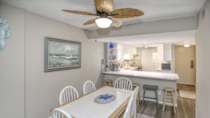A dining area with a wooden table and white chairs, a ceiling fan, and a kitchen in the background. - Oceanfront & Pool, on the Boardwalk at 37th Street (Virginia Beach)
