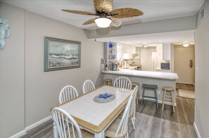 A dining area with a wooden table and white chairs, a ceiling fan, and a kitchen in the background.