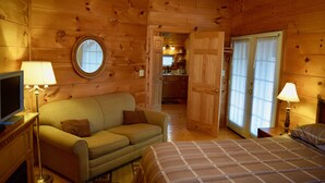 A cozy log cabin room with a bed, a sofa, a TV, and a kitchen in the background.