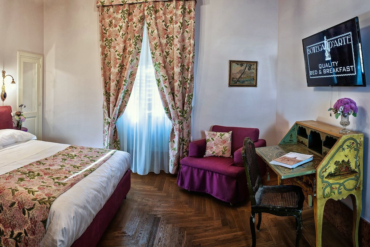 A bedroom with a large bed, a desk with a chair, a television, and a window with floral curtains.