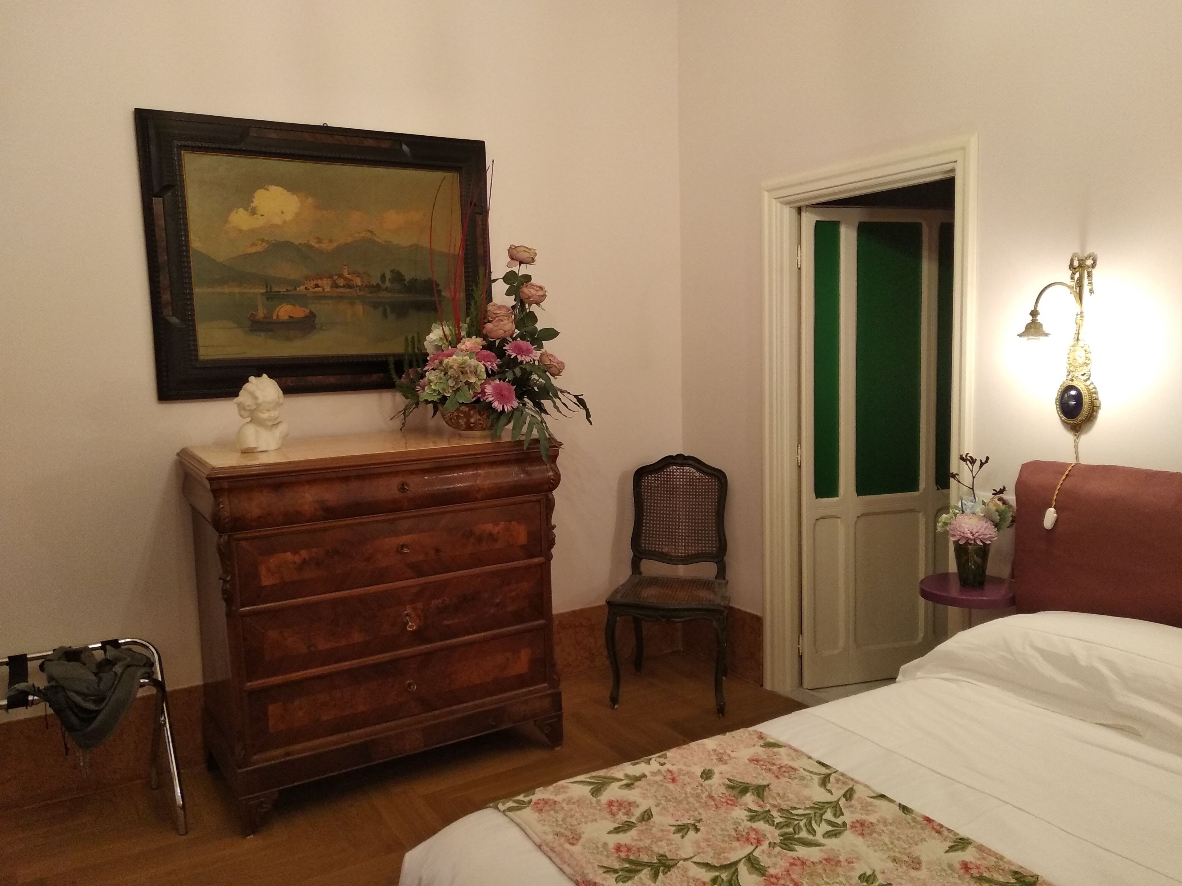 A bedroom with a wooden dresser, a chair, a bed with floral bedding, a framed painting, and a nightstand with a lamp.