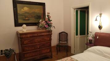 Une chambre à coucher avec une commode en bois, une chaise, un lit avec une literie à motifs floraux, un tableau encadré et une table de chevet avec une lampe.