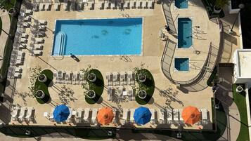 An aerial view of a resort with a large central pool, multiple smaller pools, and numerous sun loungers and umbrellas.