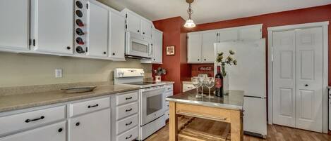 A kitchen with white cabinets, a microwave, a stove, and a refrigerator. There is a wooden table with a vase of flowers and wine glasses on it.