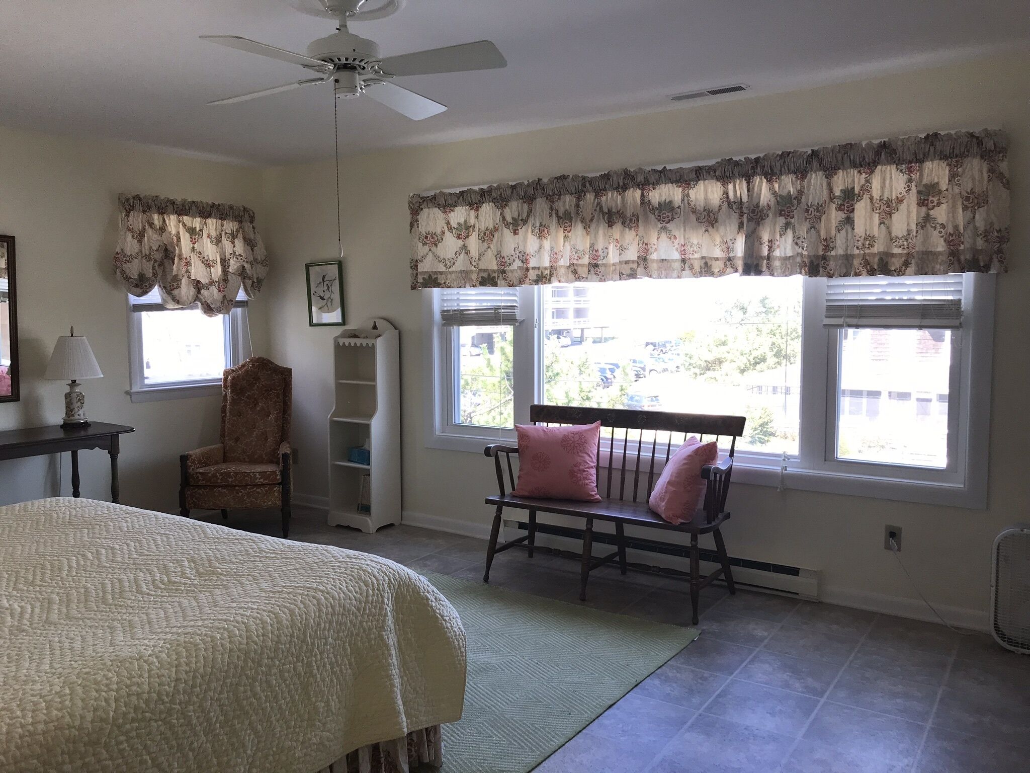Un dormitorio con cama, una silla, un ventanal con cortinas, ventilador de techo y radiador.
