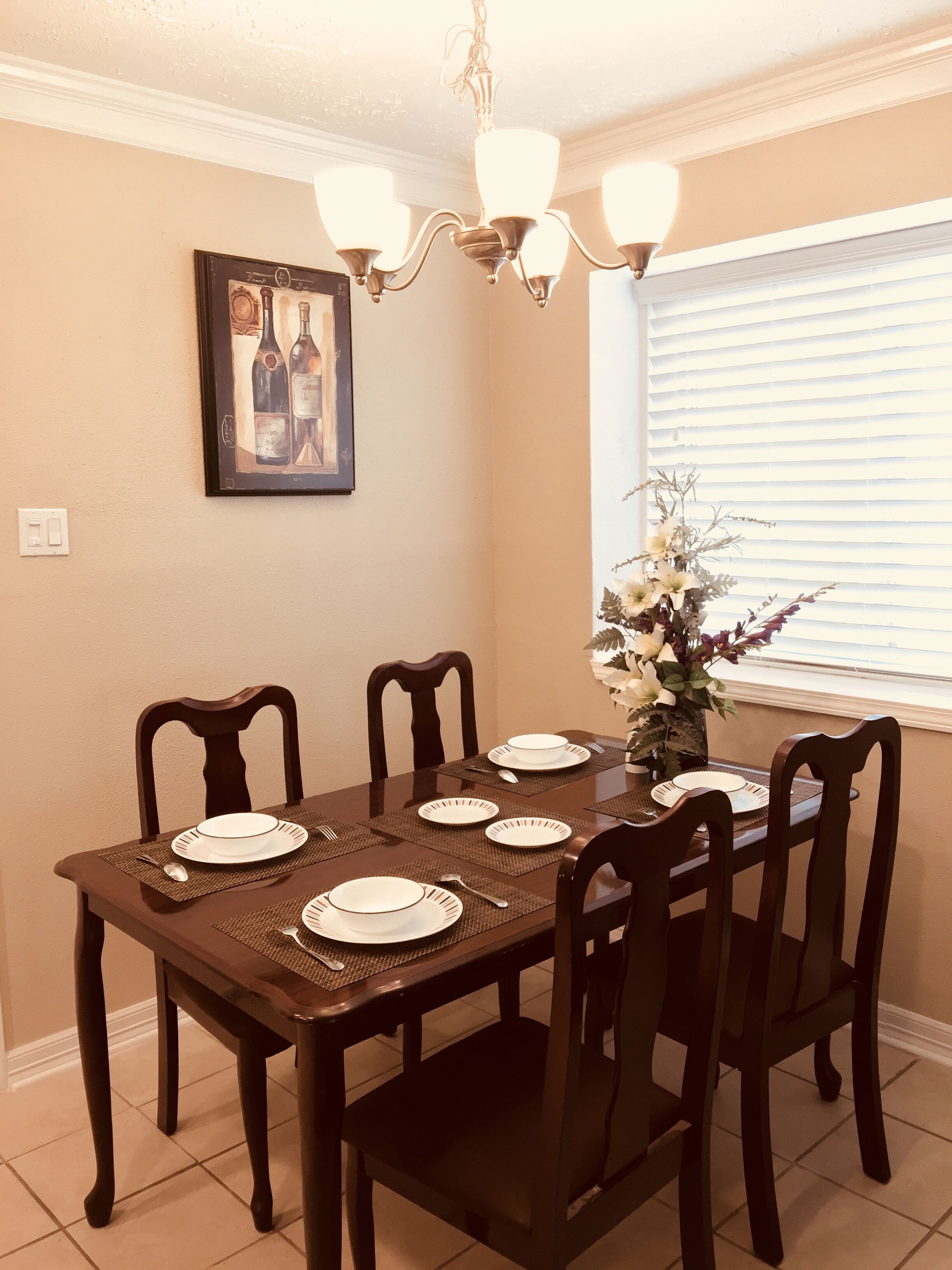 A dining room with a dark wooden table set for six, surrounded by matching chairs. A chandelier hangs from the ceiling, and a framed picture is mounted on the wall.