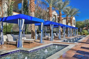 A pool area with multiple lounge chairs and umbrellas, surrounded by palm trees and a building with a tiled roof.