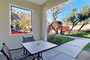 A patio with a table and chairs, a window with a metal screen, and a view of a garden with trees and shrubs.