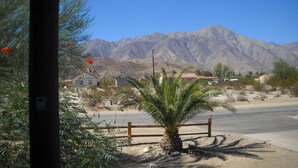 Paesaggio desertico con una catena montuosa sullo sfondo, una palma in primo piano e alcuni edifici.