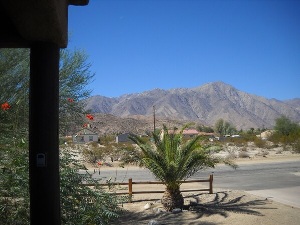 A desert landscape with a mountain range in the background, a palm tree in the foreground, and a few buildings.