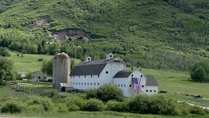Une grange blanche, ornée d’un drapeau américain, avec un silo et une maison, le tout situé dans un cadre rural, au milieu de champs et de collines verdoyantes.
