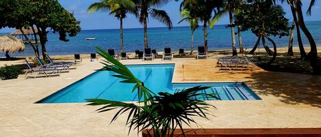 A pool area with lounge chairs, a thatched-roof structure, and palm trees.