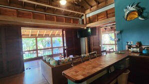 Une pièce spacieuse avec un plafond haut, des poutres en bois, une grande fenêtre, une table à manger avec des chaises, un coin cuisine et un téléviseur à écran plat fixé au mur.