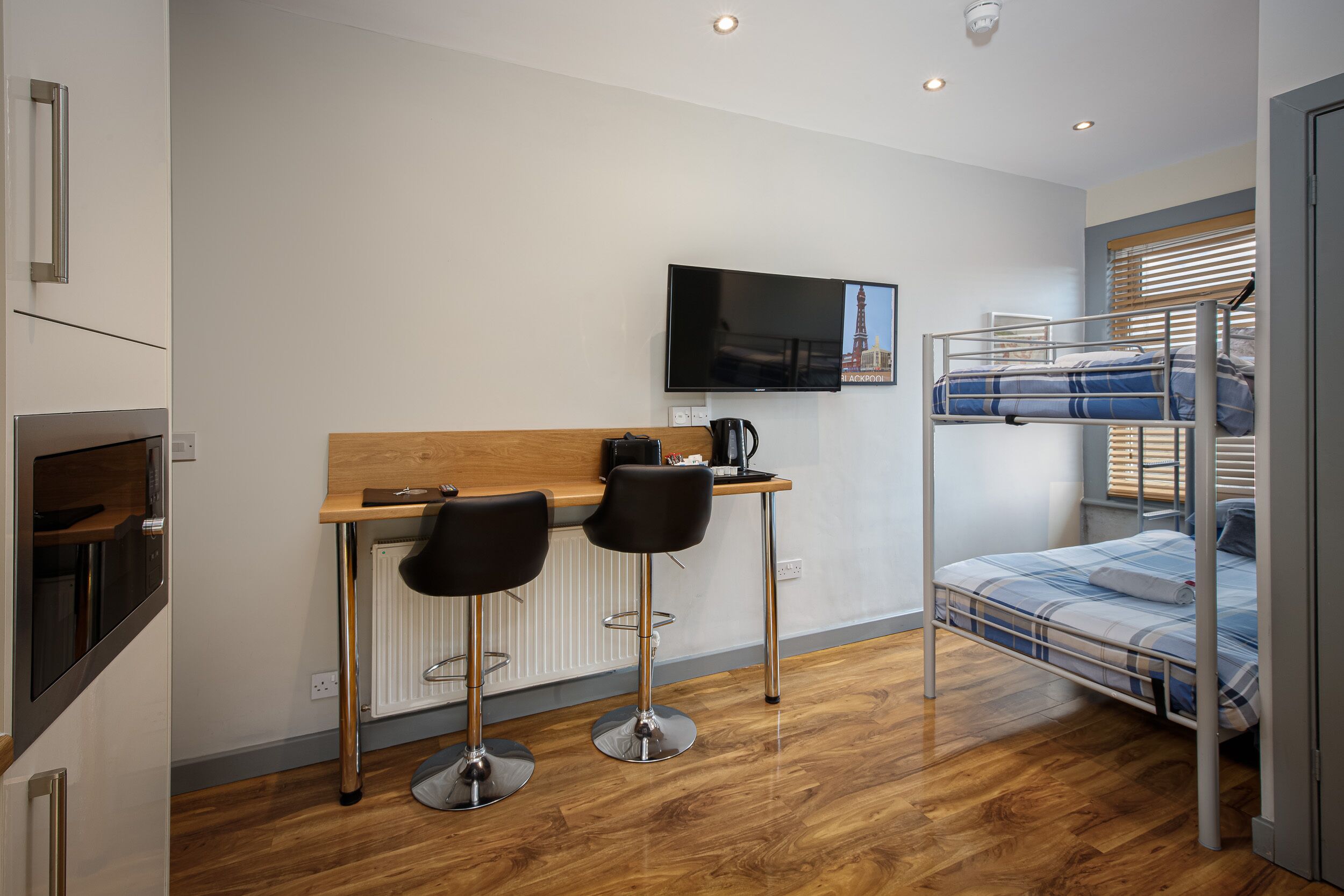 A compact room with a bunk bed, a small kitchenette, a TV mounted on the wall, and two bar stools.