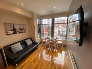 A modern living room with a black leather sofa, a dining table with chairs, large windows with blinds, and a painting on the wall.