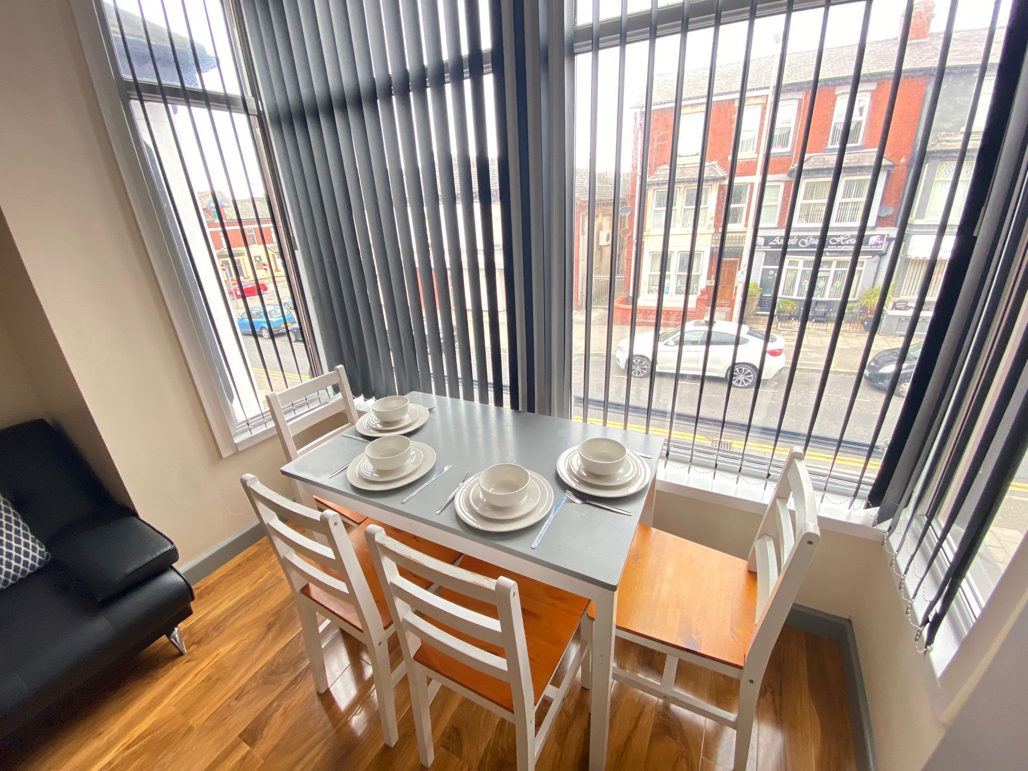 A dining area with a table set for four, white chairs, and a view of the street through large windows with blinds.