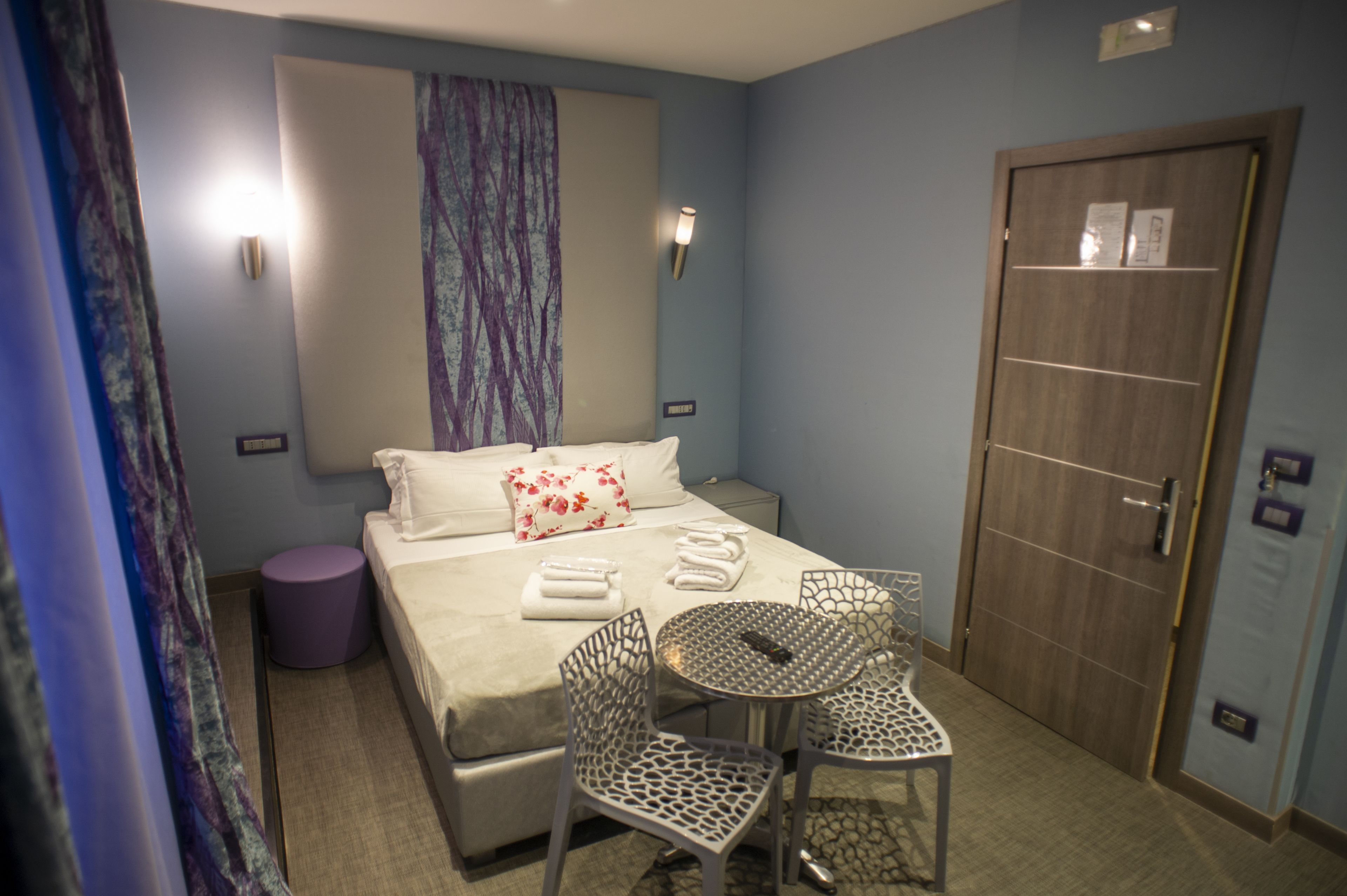 A hotel room with a bed, two chairs, a small table, a window with curtains, a door, and a wall-mounted light.