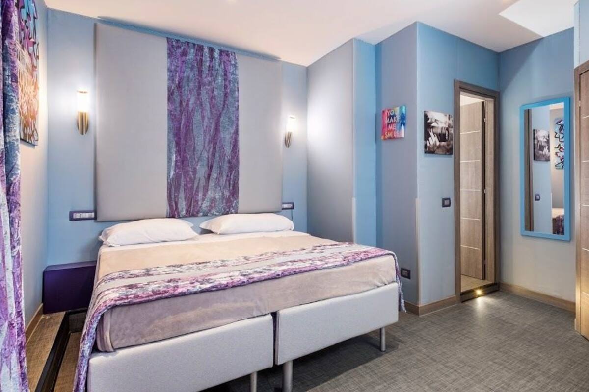 A hotel room with a bed, a nightstand, a wall-mounted lamp, a door, and a mirror.