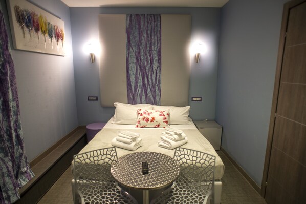 Una habitación de hotel con una cama, dos lámparas de noche, una mesa redonda con sillas y una mesita auxiliar.