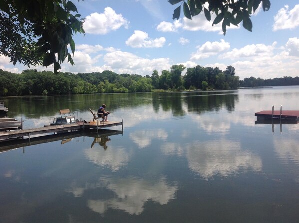 A calm lake with a dock, a person sitting on a chair, and a boat.