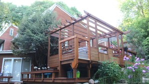 A wooden deck with a staircase leading to a covered area, surrounded by greenery and a stone wall.