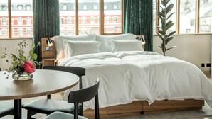 Un dormitorio moderno con una cama grande, una mesa redonda con sillas y vistas a edificios a través de amplios ventanales.