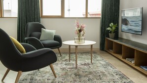 Una sala de estar moderna con dos sillones negros, una mesita redonda con un florero y un televisor en la pared, además de una alfombra estampada.