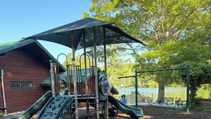A playground with a slide and climbing structures, surrounded by trees and a building. - Nanny’s River Cabin_Peaceful Blue Ridge Mountain Retreat in Linville Land Harbor (Newland)